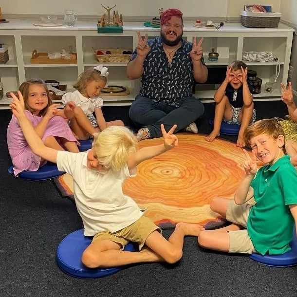 Children sitting in a circle with an adult on a colorful rug in a classroom setting, smiling and making peace signs, possibly in an Episcopal church s