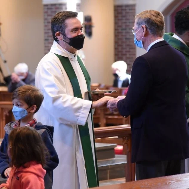 Priest in white robe and mask greets masked congregant at an Episcopal church service, with children nearby.