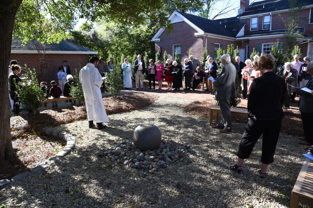 Gathering in an episcopal church garden with a minister leading, surrounded by attendees holding programs, near a stone sphere centerpiece.